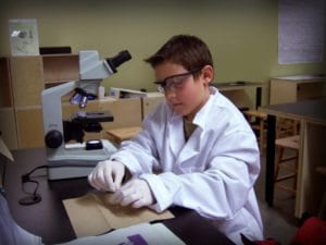 Child in lab coat looking at a slide under a microscope.