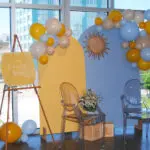 A yellow and blue baby shower photo booth backdrop with varying sizes of yellow, blue, and yellow balloons with a decorative golden sun. A sign reads "Here comes the son"
